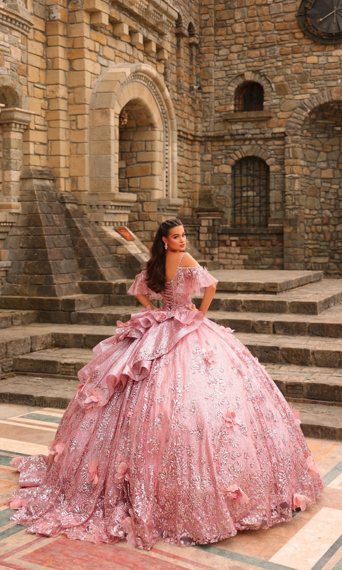 A woman in the Amarra 54229 Glitter Tulle Quinceañera Dress stands on stone steps before a grand, ornate stone building.